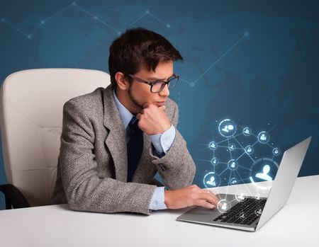Attractive young man sitting at desk and typing on laptop with social network icons comming out