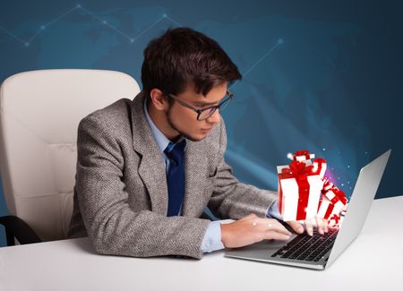 Handsome young man sitting at desk and typing on laptop with present boxes icons