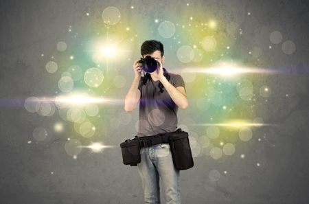 A young amateur photographer with professional camera equipment taking picture in front of grey wall full of colorful bokeh and glowing lights concept