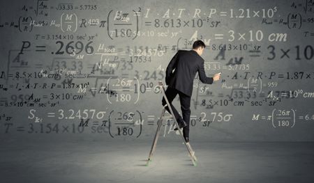 A businessman in elegant suit standing on a small ladder and writing numbers, calculating on grey wall background