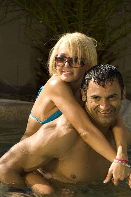 Pretty Caucasian blond in bikini hangs on to smiling Hispanic man in backyard swimming pool