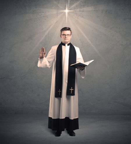 A young male priest in black and white giving his blessing in front of grey wall with glowing cross concept.