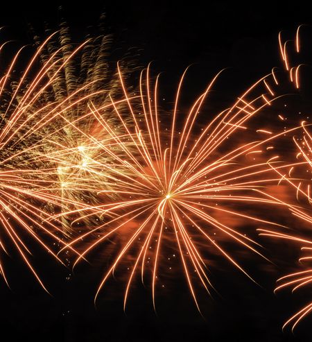 Bursts of yellowish fireworks near puffs of reddish smoke and motion-blurred streaks from previous explosion