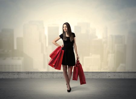 Attractive lady in black holding red shopping bags standing in front o urban landscape with tall buildings concept