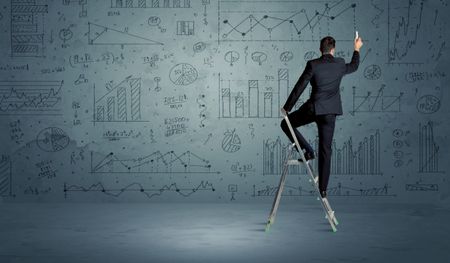 A businessman in modern stylish elegant suit standing on a small ladder and drawing pie and block charts on grey wall background with exponential progressing curves, lines, circles,  blocks, numbers