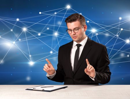 Young handsome businessman sitting at a desk with a blue connection graphic behnid him 