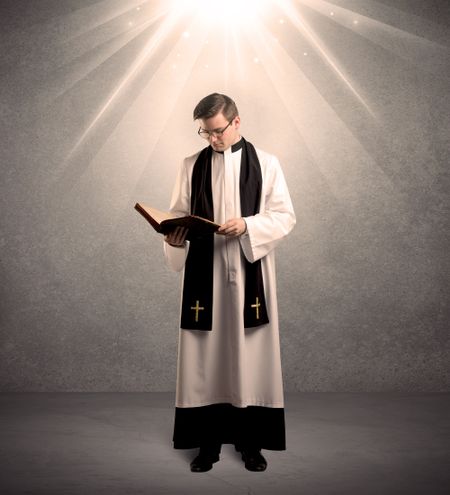 A male religious young priest in black and white dress giving his blessing, holding the holy bible while being illuminated from strong light beams coming from above concept
