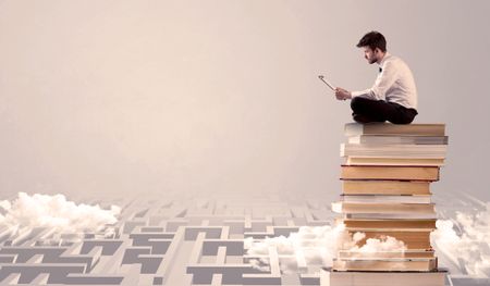 A businessman with laptop tablet in elegant suit sitting on a stack of books on top of sandy labirynth background concept