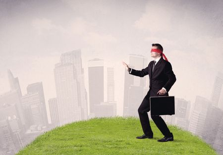 Young blindfolded businessman steps on a a patch of grass with a cloudy city in the background