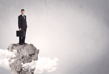 A happy successful businessman standing on a stone cliff with clouds in front of clear empty grey background concept