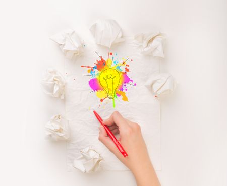 Female hand next to a few crumpled paper balls drawing a colorful lightbulb