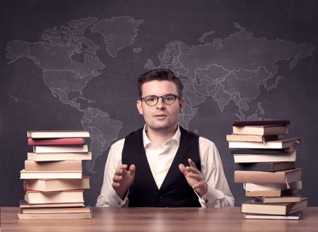A young ambitious geography teacher in glasses sitting at classroom desk with pile of books in front of world map drawing on blackboard, back to school concept.