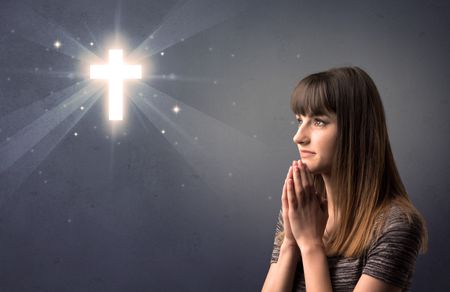 Young woman praying on a grey background with a shiny cross above her