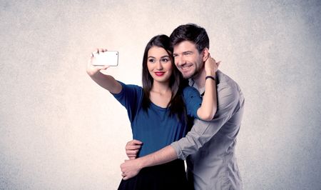 A young couple in love taking selfie with a mobile phone in the handsome guy's hand in front of an empty clear grey wall background concept