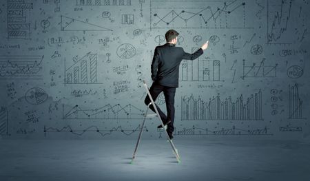 A businessman in modern stylish elegant suit standing on a small ladder and drawing pie and block charts on grey wall background with exponential progressing  lines, circles, angles, blocks, numbers