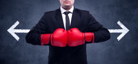 A confident businessman standing with red boxing gloves on his hand in front of arrows pointing in different directions on urban wall background concept.